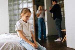 Porträt einer depressiven süßen kleinen Tochter, die traurig auf der Couch sitzt, während die Eltern im Wohnzimmer im Hintergrund streiten und kämpfen, Konzept von Familienproblemen.