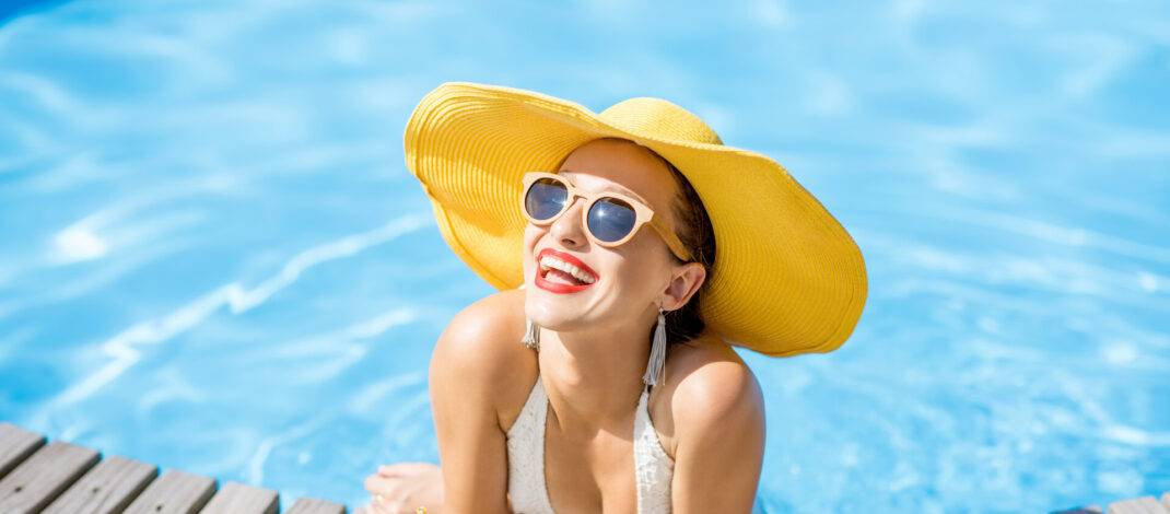 Junge Frau im Badeanzug mit großem gelben Sonnenhut entspannt sich mit einem frischen Getränk am Pool