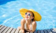 Junge Frau im Badeanzug mit großem gelben Sonnenhut entspannt sich mit einem frischen Getränk am Pool