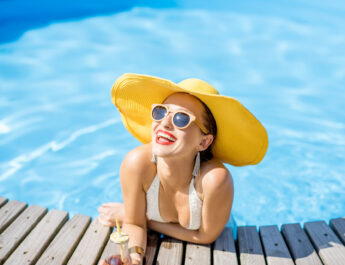 Junge Frau im Badeanzug mit großem gelben Sonnenhut entspannt sich mit einem frischen Getränk am Pool