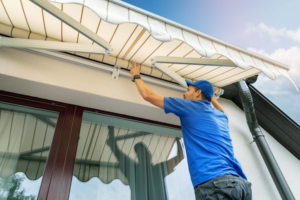 Ein Fachmann in blauer Arbeitskleidung installiert eine Markise an der Außenwand eines Hauses, um optimalen Sonnenschutz für Fenster und Terrasse zu gewährleisten.
