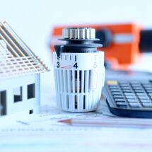 Ein Thermostat eingestellt auf eine energieeffiziente Temperatur neben einem Modellhaus, einem Bauhelm und einem Taschenrechner, symbolisch für die Überwachung und Optimierung der Heizölpreise in Sachsen.