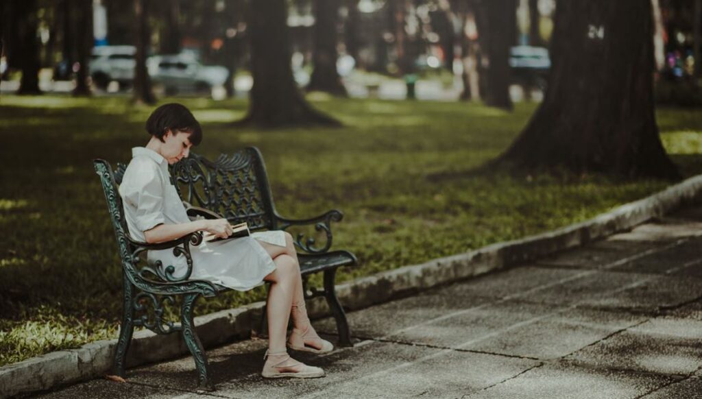 Eine Frau sitzt auf einer Parkbank in einem grünen Park in Frankfurt und liest ein Buch, umgeben von Bäumen und Natur.