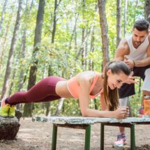 Eine Frau macht unter Anleitung ihres Personal Trainers eine Plank-Übung auf einer Bank im Kölner Stadtwald. Die grüne Umgebung bietet eine ruhige und motivierende Atmosphäre für ein intensives Outdoor-Training.