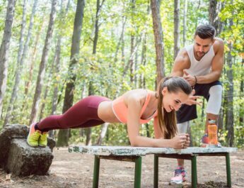 Eine Frau macht unter Anleitung ihres Personal Trainers eine Plank-Übung auf einer Bank im Kölner Stadtwald. Die grüne Umgebung bietet eine ruhige und motivierende Atmosphäre für ein intensives Outdoor-Training.