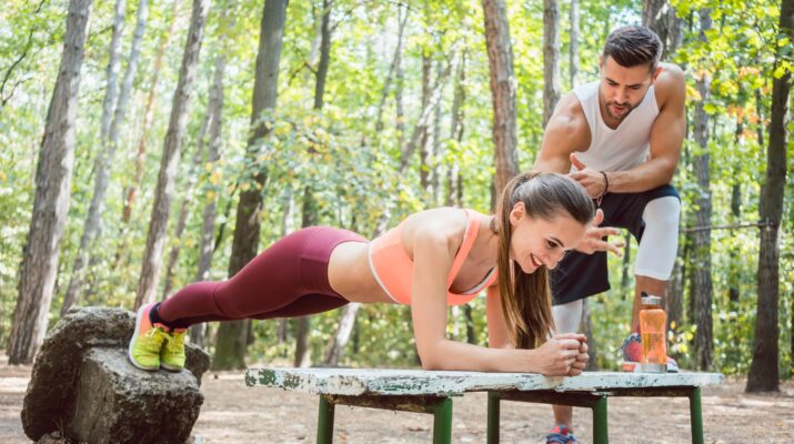 Eine Frau macht unter Anleitung ihres Personal Trainers eine Plank-Übung auf einer Bank im Kölner Stadtwald. Die grüne Umgebung bietet eine ruhige und motivierende Atmosphäre für ein intensives Outdoor-Training.