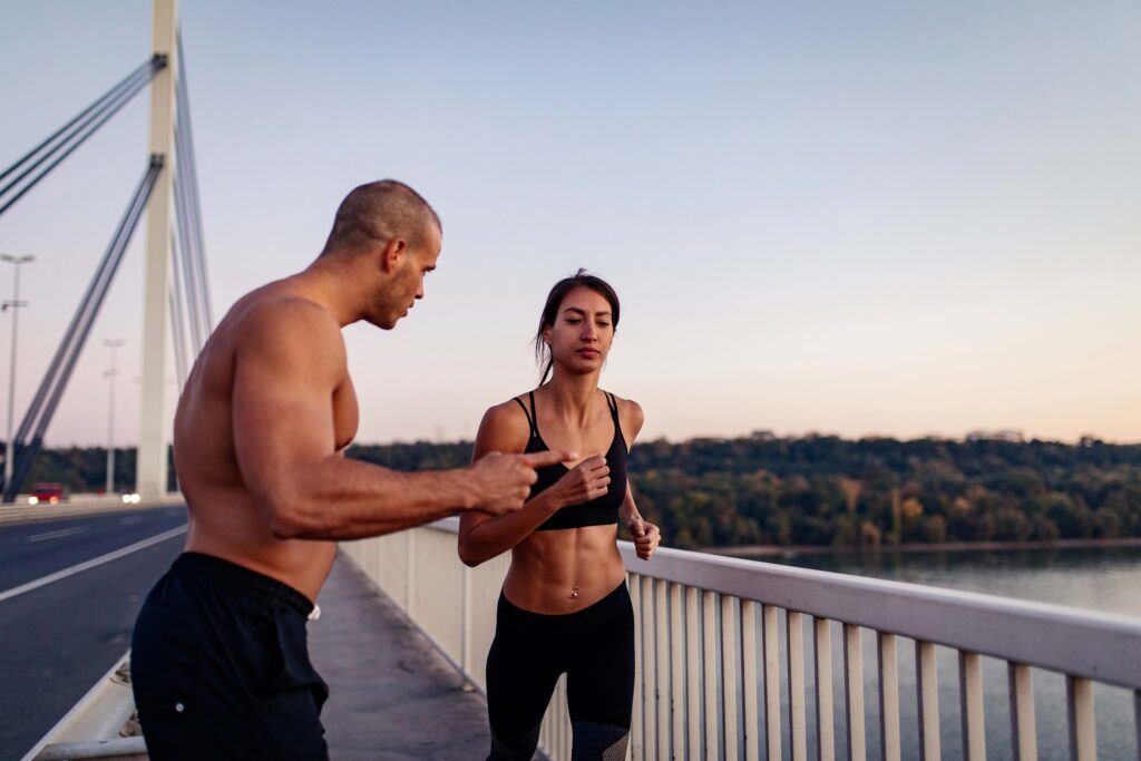 Ein Personal Trainer gibt einer Frau beim Lauftraining auf der Severinsbrücke in Köln Anweisungen. Die Morgenstimmung und der Blick auf den Rhein schaffen eine inspirierende Umgebung für ein intensives Cardio-Workout.