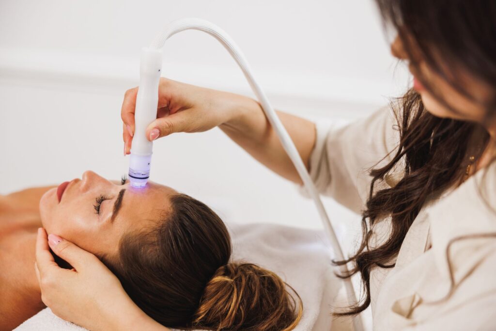 Eine Frau erhält eine Hydrafacial Behandlung im Gesicht, während eine Kosmetikerin das Gerät sanft über die Stirn führt