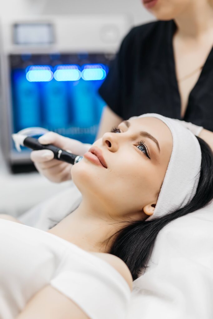 Eine Frau erhält eine Hydrafacial Gesichtsbehandlung mit modernen Geräten, während sie entspannt auf einer Liege liegt und von einer Kosmetikerin betreut wird