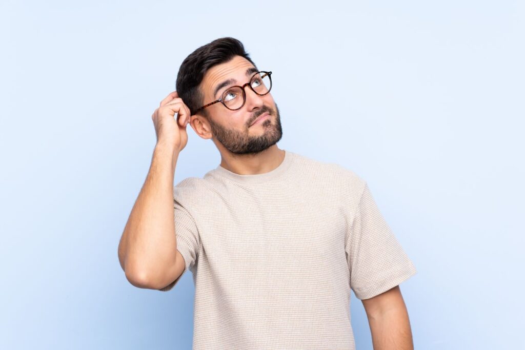 Nachdenklicher Mann mit Brille kratzt sich am Kopf und schaut nach oben vor einem hellblauen Hintergrund