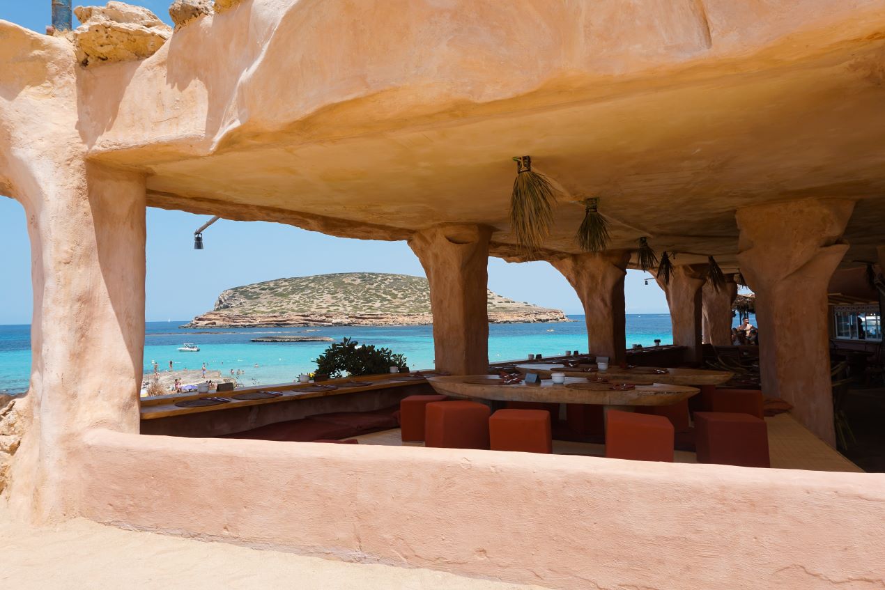 Ein stilvolles Ibiza Restaurant an der Cala Conta mit offenem Design, gemütlichen Sitzgelegenheiten und einem Panoramablick auf das türkisfarbene Wasser und die vorgelagerte Insel.