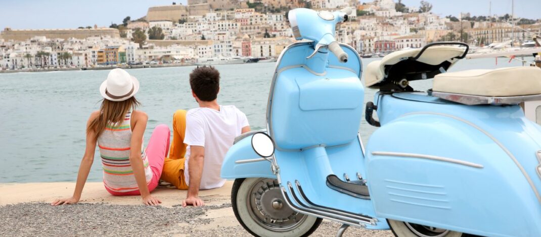 Ein Paar sitzt am Hafen und blickt auf die Altstadt von Ibiza-Stadt, mit einer blauen Vespa im Vordergrund und Booten im Hintergrund.