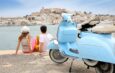 Ein Paar sitzt am Hafen und blickt auf die Altstadt von Ibiza-Stadt, mit einer blauen Vespa im Vordergrund und Booten im Hintergrund.