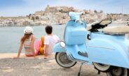 Ein Paar sitzt am Hafen und blickt auf die Altstadt von Ibiza-Stadt, mit einer blauen Vespa im Vordergrund und Booten im Hintergrund.