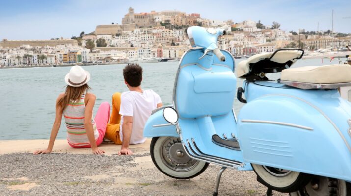 Ein Paar sitzt am Hafen und blickt auf die Altstadt von Ibiza-Stadt, mit einer blauen Vespa im Vordergrund und Booten im Hintergrund.