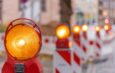 Reihe von gelben Baustellenleuchten mit Warnbaken auf städtischer Straße bei Tageslicht, Fokus auf Vordergrundleuchte