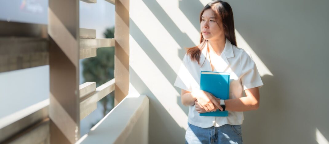 Junge Frau steht im Sonnenlicht mit Heft in der Hand und schaut nachdenklich aus dem Fenster – Symbol für Neuanfang und berufliche Orientierung