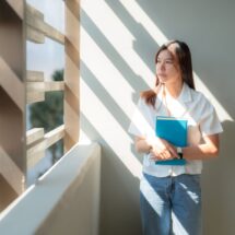 Junge Frau steht im Sonnenlicht mit Heft in der Hand und schaut nachdenklich aus dem Fenster – Symbol für Neuanfang und berufliche Orientierung