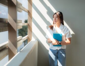 Junge Frau steht im Sonnenlicht mit Heft in der Hand und schaut nachdenklich aus dem Fenster – Symbol für Neuanfang und berufliche Orientierung