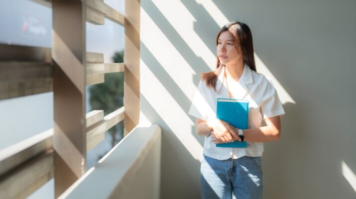 Junge Frau steht im Sonnenlicht mit Heft in der Hand und schaut nachdenklich aus dem Fenster – Symbol für Neuanfang und berufliche Orientierung